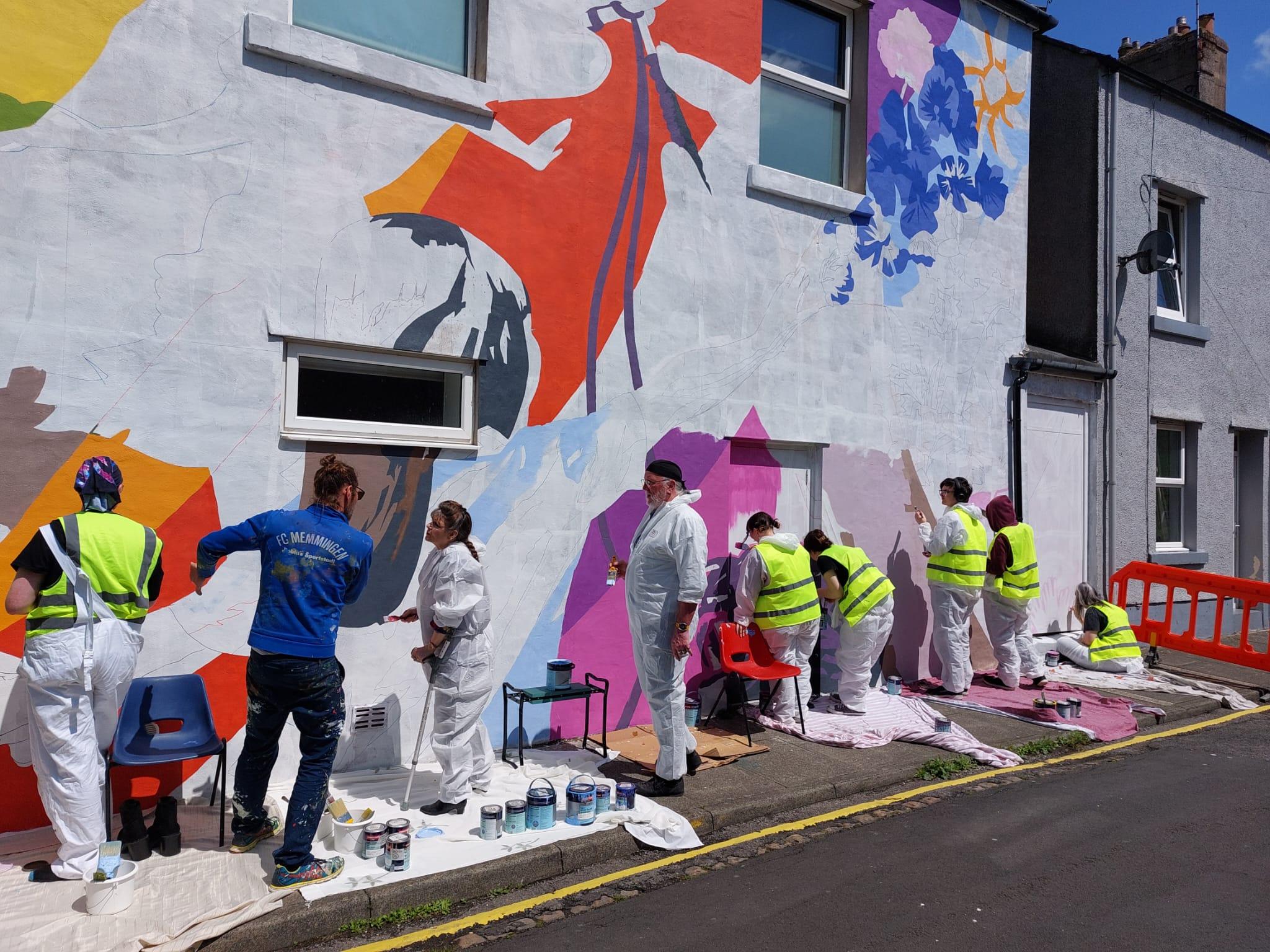 A row of people in white overalls and yellow hi-vis vests painting a brightly coloured mural.