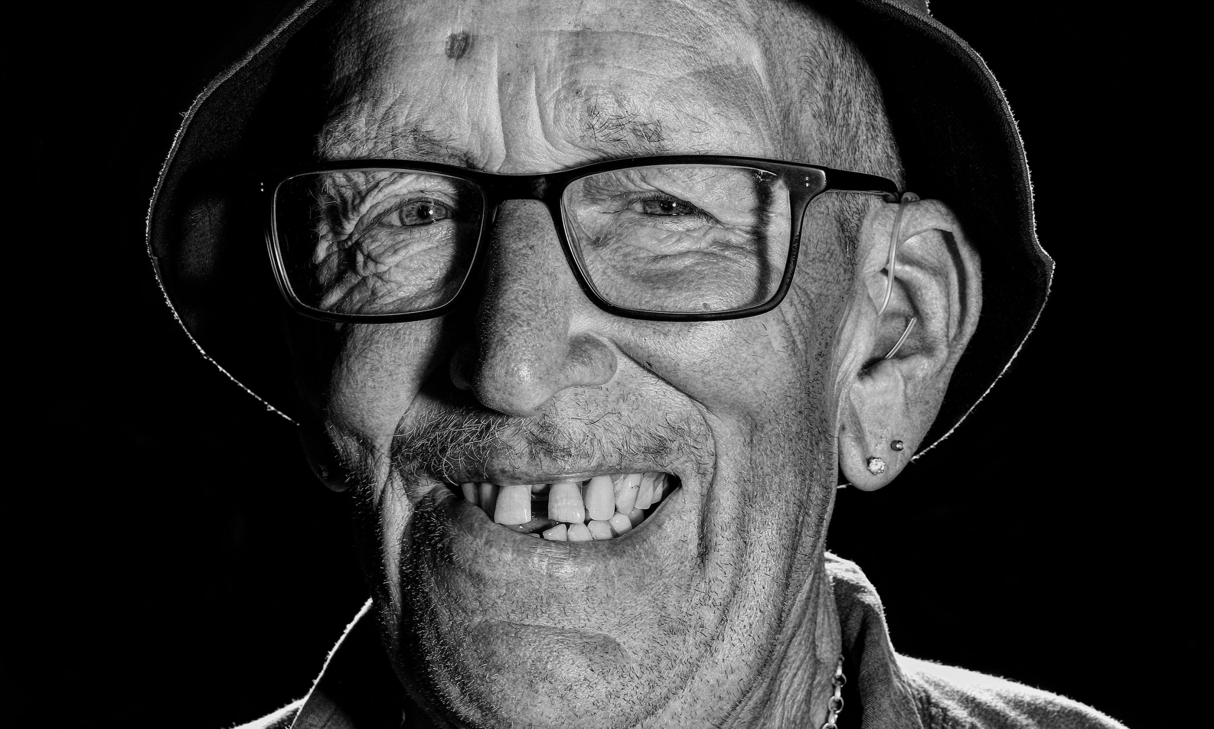 A black and white portrait of an older white man smiling wearing glasses and a Guinness bucket hat.